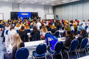 Students attending Beacon 2023. Students sit at fold-out tables and cushioned chairs face a stage decorated with the Beacon logo.